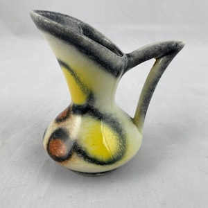 Pode incluir: Um pequeno jarro de cerâmica vintage com um design único. O jarro apresenta uma base creme com padrões abstratos amarelos, pretos e castanhos. Possui uma alça curva e um bico com uma borda cinza.