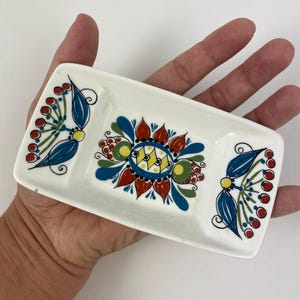 Op de afbeelding: Een rechthoekige, witte keramische schaal met drie compartimenten. Het heeft een kleurrijk bloemmotief in rood, blauw, geel en groen. De schaal wordt in de hand van een persoon gehouden.