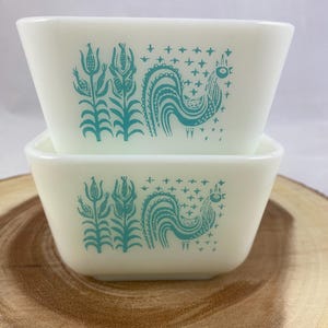 Peut inclure: Deux bols carrés Pyrex blanc cassé superposés. Chaque bol présente un coq et un motif floral turquoise. Les bols sont posés sur une surface en bois.