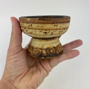 May include: A small, handcrafted ceramic bowl with a pedestal base. The bowl is light brown and tan with dark brown accents. The pedestal base has a spiral design. The bowl is held in a hand.
