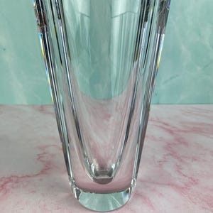 Op de afbeelding: Een heldere, facetgeslepen glazen vaas met een brede, gebogen opening en een smalle basis. De vaas heeft verticale groeven en een glad, gepolijst oppervlak. De achtergrond is een mix van roze en teal.
