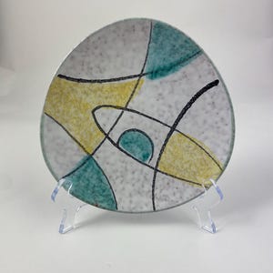 Peut inclure: Petite assiette triangulaire en céramique au design moderne du milieu du siècle. L'assiette présente un fond blanc moucheté avec des formes abstraites turquoise et jaunes, et des accents linéaires noirs. L'assiette est présentée sur un présentoir en plastique transparent.