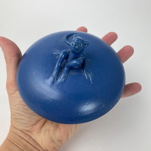 Op de afbeelding: Een ronde, kobaltblauwe keramische deksel met een gebeeldhouwde apenfiguur. De deksel heeft een matte afwerking. De aap is ook blauw en is aan de deksel bevestigd. De deksel is ongeveer 10 cm in diameter.