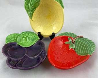 May include: Three ceramic fruit-shaped dishes: a yellow lemon, a purple blackberry, and a red strawberry. Each dish has green leaves and a glossy finish. The lemon dish is the largest, with a brown stem.