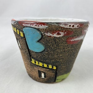 Puede incluir: Un pequeño macetero de cerámica marrón con un diseño abstracto colorido. El macetero tiene un borde blanco y presenta un diseño azul, amarillo, rojo y verde.