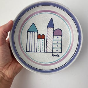 May include: A ceramic plate with a hand-painted city scene. The plate features a white background with a blue, pink, and teal ringed border. The city scene includes buildings with blue, red, and purple accents. The artist's initials, "kmk", are visible.