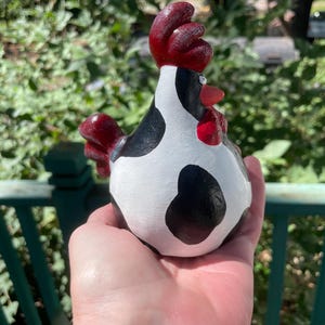 May include: A hand holds a black and white ceramic chicken figurine with a red comb and wattle. The chicken is painted to look like a cow with black spots on a white background.