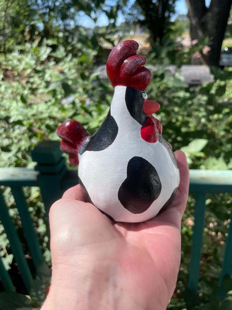 May include: A hand holds a black and white ceramic chicken figurine with a red comb and wattle. The chicken is painted to look like a cow with black spots on a white background.