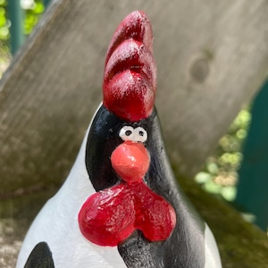 May include: A hand-painted ceramic chicken figurine with black and white spots and a red comb. The chicken has a whimsical expression with large eyes and a red beak.