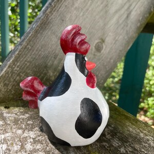 May include: A black and white cow-patterned ceramic rooster figurine with a red comb and wattle. The rooster is standing on a wooden surface.