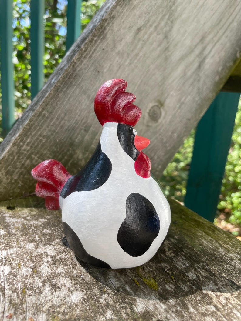 May include: A black and white cow-patterned ceramic rooster figurine with a red comb and wattle. The rooster is standing on a wooden surface.