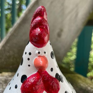 May include: A white ceramic chicken figurine with black polka dots. The chicken has a red comb and wattle, and a pink beak.