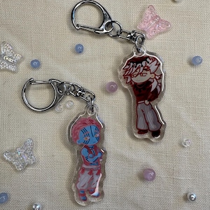 May include: Two anime-style character keychains with silver hardware. One keychain features a character with red hair and a dark red outfit. The other keychain has a blue-skinned character with blue and red clothing. Decorative beads and butterfly charms surround the keychains.