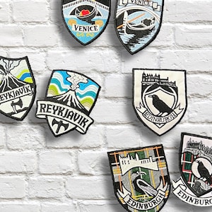 May include: Embroidered shield patches with city-themed designs. The patches display names such as Paris, Venice, Reykjavik, and Edinburgh. Each patch showcases distinct illustrations and colour schemes, including a volcano, a raven, and architectural details.