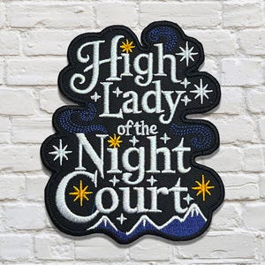 Puede incluir: Parche bordado con el texto "High Lady of the Night Court" en escritura blanca. El parche tiene un fondo negro con detalles en azul y dorado, incluyendo estrellas y detalles de montañas. El parche está sobre una pared de ladrillos blancos.