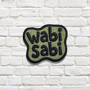 May include: Embroidered patch with the words "wabi sabi" in black, on a green background, with a black border. The patch is against a white brick wall.