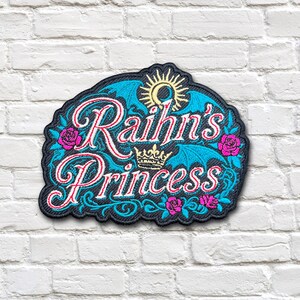 Puede incluir: Un parche bordado con el texto Raihn's Princess en letra blanca con contornos rosas. El diseño incluye una corona dorada, un sol amarillo, alas de murciélago azules y rosas de color rosa sobre un fondo negro con un borde festoneado.
