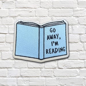 May include: A light blue embroidered patch shaped like an open book with black stitched borders. The right page features the black embroidered text "GO AWAY, I'M READING" in a bold, sans-serif font. The patch has a white edge border.
