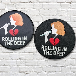 May include: Two black circular patches featuring a singer silhouette, a broken heart, and the text "ROLLING IN THE DEEP" in white. The singer has orange hair and is holding a microphone. The patches are on a white brick wall.