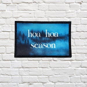 May include: A rectangular patch with a black border and a blue background featuring a forest silhouette. The words "hoa hoa season" are written in white, stylized font across the center of the patch.