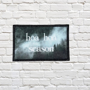 May include: A rectangular patch with a black border, featuring the words "hoa hoa season" in white, stylized text. The background shows a misty forest scene in shades of gray and green, set against a white brick wall.