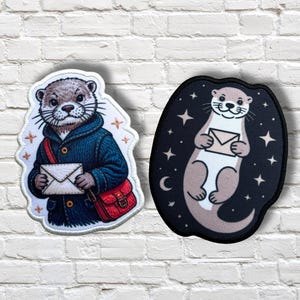 May include: Two embroidered patches featuring otters. One otter wears a blue coat and red messenger bag, holding an envelope. The other otter is light brown with a dark background, holding an envelope, surrounded by stars and a crescent moon.