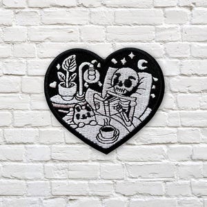 May include: A heart-shaped patch with a black border. The design shows a skeleton reading in bed, with a plant, cat, lamp, and coffee cup in white against a black backdrop. The patch is displayed against a white brick wall.