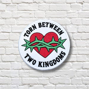 May include: A round, white patch with the text "TORN BETWEEN TWO KINGDOMS" in black. The patch features a red heart pierced by a green thorn crown.