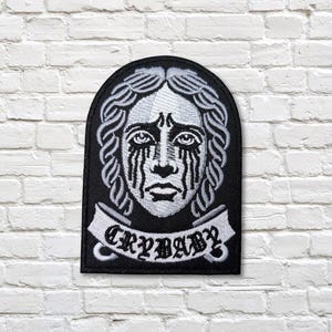 May include: Black and white embroidered patch featuring a stylized crying face with tears. The patch has a banner at the bottom with the word "CRYBABY" in white lettering. The patch is rectangular with an arched top.