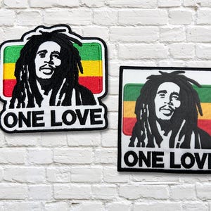 Puede incluir: Dos parches bordados con un retrato en blanco y negro con rastas, sobre un fondo verde, amarillo y rojo. El texto "ONE LOVE" está debajo del retrato. Un parche tiene un borde blanco, el otro un borde negro.