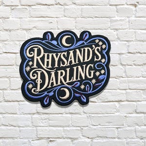 Peut inclure: Écusson brodé avec le texte "Rhysand's Darling" dans une police stylisée. Le patch présente un fond noir avec des accents bleus et blancs, y compris des motifs floraux et étoilés. L'écusson est contre un mur de briques blanches.