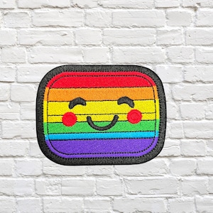 Pode incluir: Patch bordado com um rosto sorridente em um design de arco-íris. O patch tem uma borda preta e inclui as cores vermelho, laranja, amarelo, verde, azul e roxo. O design está em uma parede de tijolos brancos.