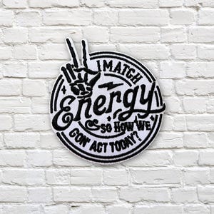 May include: A circular black and white patch featuring the text "I match energy so how we gon' act today?" and a peace sign hand gesture. The patch is on a white brick wall.