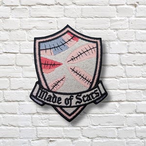 May include: Embroidered patch in a shield shape with a black border. The patch features a light grey background with pink, blue, and red stitched designs. The text "Made of Scars" is displayed on a banner at the bottom.