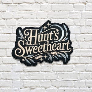 May include: Embroidered patch with the words "Hunt's Sweetheart" in elegant script. The patch is black with cream-coloured lettering and decorative floral and celestial accents. The patch is set against a white brick wall.