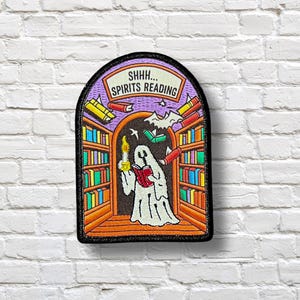 Puede incluir: Parche bordado que representa un fantasma leyendo en una biblioteca. El parche tiene un borde negro y el texto "SHHH... SPIRITS READING". El fantasma sostiene una vela y un libro, rodeado de libros flotantes y un murciélago.