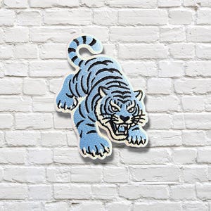 Könnte beinhalten: Ein gestickter Aufnäher, der einen hellblauen Tiger mit dunkelblauen Streifen in Sprungposition zeigt. Der Tiger hat ein geöffnetes Maul mit sichtbaren Zähnen, spitze Ohren und einen geschwungenen Schwanz. Das Motiv ist weiß umrandet auf strukturiertem Grund.