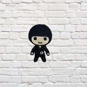 Puede incluir: Parche bordado con un personaje de dibujos animados con un atuendo negro y gorra a juego. El personaje tiene una cara sonriente y está sobre una pared de ladrillos blancos. Ideal para ropa o accesorios.