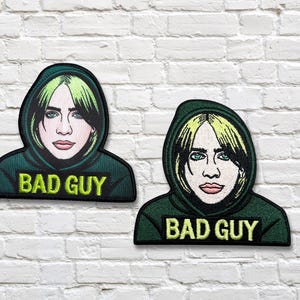 以下が含まれることがあります： 緑と黒の髪の人物が緑のパーカーを着ている肖像画が描かれた2つの刺繍パッチ。「BAD GUY」の文字がパーカーの前面に刺繍されています。パッチは白いレンガの壁にあります。