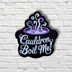 May include: Embroidered patch featuring a black cauldron with purple liquid and steam. The cauldron has a purple rim and the text "Cauldron Boil Me!" in white script. The patch is set against a white brick wall.