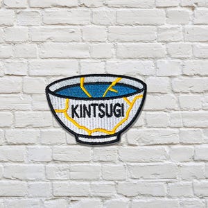 Könnte beinhalten: Gestickter Aufnäher mit einer weißen Schüssel mit blauem Innenraum und goldfarbenen Rissen, mit dem Wort "KINTSUGI" in schwarzen Buchstaben. Der Aufnäher ist schwarz umrandet und hat einen weißen Rand. Der Aufnäher wird vor einer weißen Backsteinmauer gezeigt.
