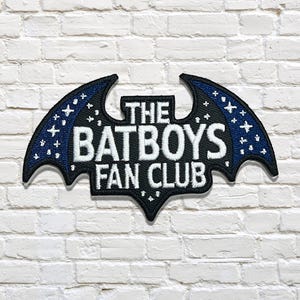 May include: A black and blue embroidered patch in the shape of a bat with the words "THE BATBOYS FAN CLUB" in white. The bat wings have a blue background with white star and cross accents. The patch is on a white brick wall.