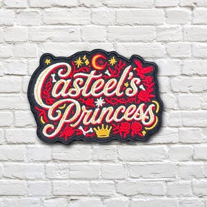 May include: An embroidered patch with a black background featuring the text Casteel's Princess in white script with yellow outlines. The design includes red roses, thorny vines, a yellow crown, a red crescent moon, small stars, and white bones.