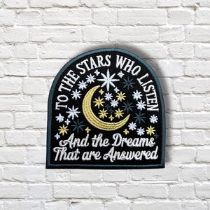 May include: Embroidered patch with a black background and a crescent moon and stars design. The patch reads "TO THE STARS WHO LISTEN" and "And the Dreams That are Answered" in white lettering. The patch is arched at the top.