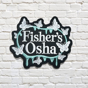 Könnte beinhalten: Schwarzer, gestickter Aufnäher mit dem weißen Text "Fisher's Osha", umgeben von einem türkisfarbenen Rand und weißen Schmetterlingen. Der Aufnäher befindet sich auf einer weißen Backsteinmauer.