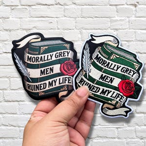 May include: Two embroidered patches featuring a dark green book with gold accents, a red rose, and a white quill. A white banner wraps around the book with the black text "MORALLY GREY MEN RUINED MY LIFE". One patch has a black border, the other a white border.