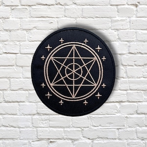 May include: A black, circular embroidered patch featuring a gold-colored geometric design. The design includes a central circle, a six-pointed star, and surrounding cross-shaped symbols. The patch is likely for clothing or accessories.