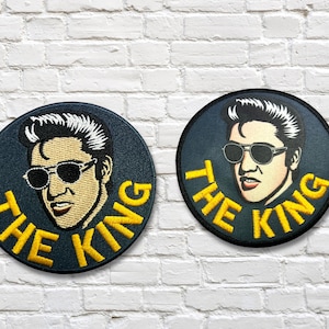 May include: Two circular patches featuring a portrait of Elvis Presley in sunglasses. The patches have a dark blue background with yellow text that reads "THE KING". The patches are on a white brick wall.