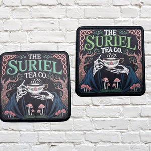 Pode incluir: Dois patches quadrados com o texto "THE SURIEL TEA CO." em verde e branco. Os patches apresentam um esqueleto segurando uma xícara de chá com um desenho floral, rodeado por cogumelos e árvores. O fundo é preto.