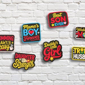 May include: A collection of colorful patches with text, including "Momming Ain't Easy," "Mama's Boy Forever," "Best Son Ever," and "World's Best Mom." The patches are on a white brick wall.